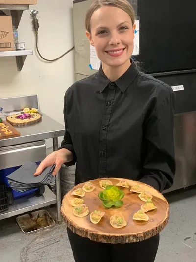 Smiling server holding a board of avocado-topped canapés in a kitchen